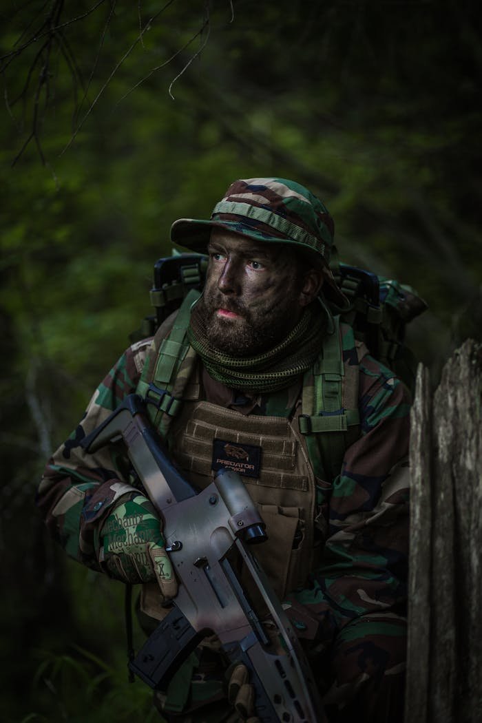 Bearded soldier in camouflage uniform with rifle in dense forest setting.
